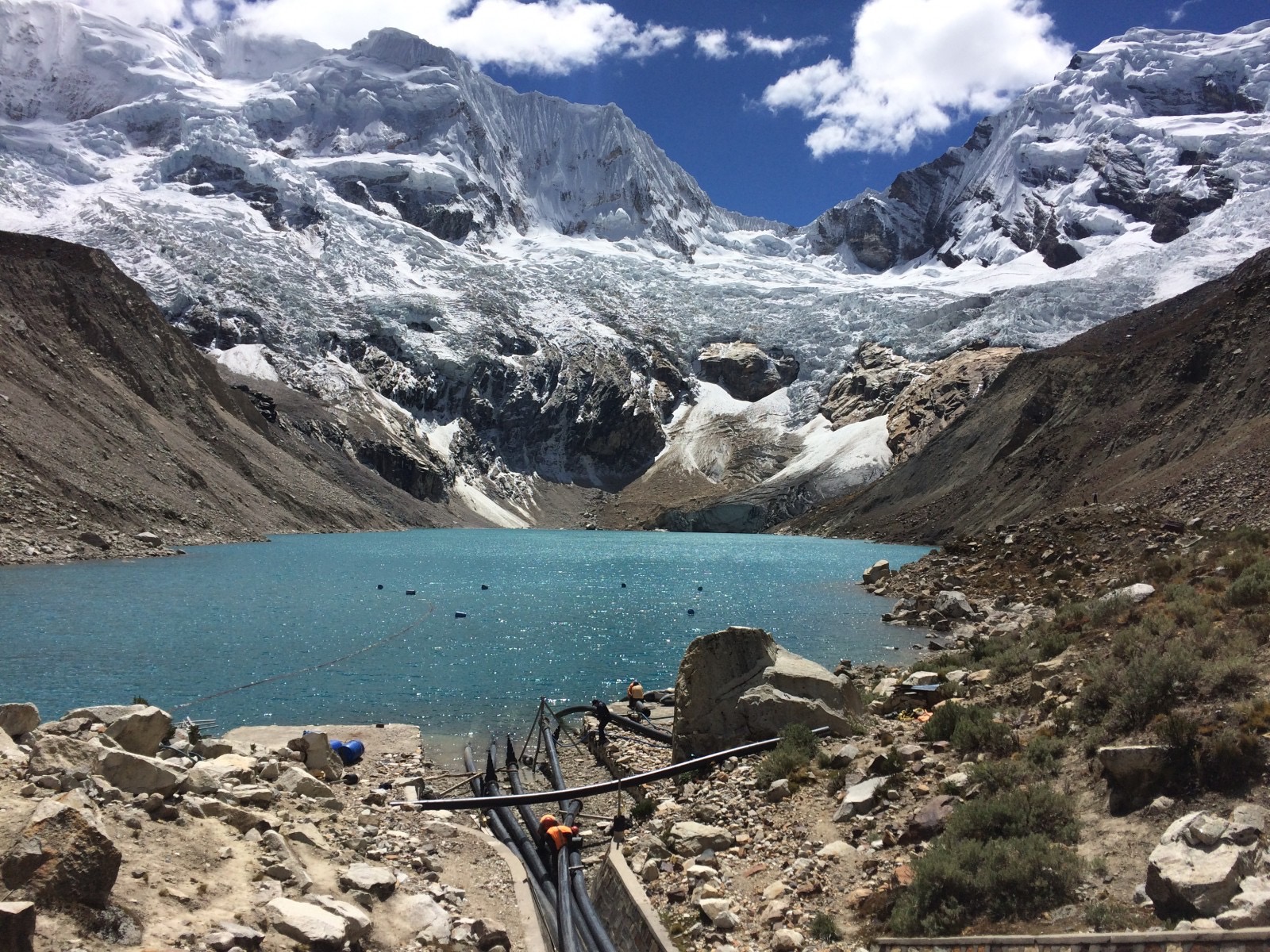 A glacier lake with visible human intervention infrastructure and drainage pipes in the foreground